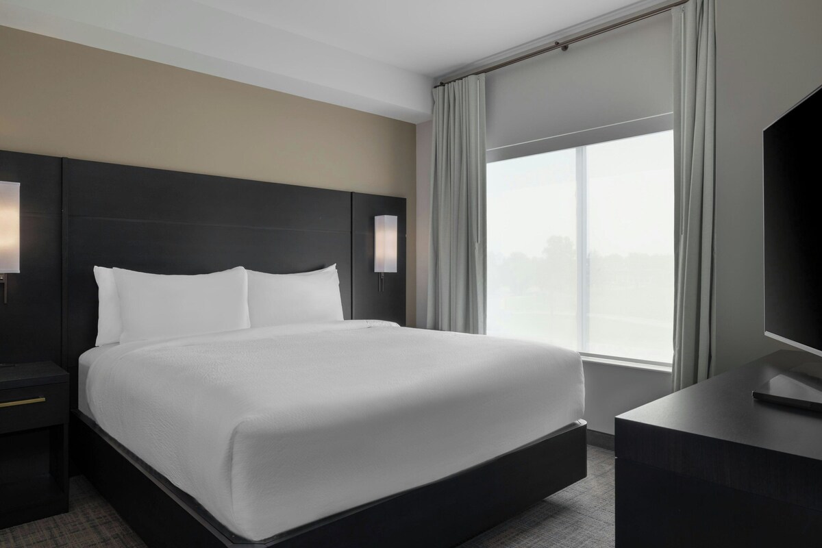 Habitación de hotel con una cama grande, televisión de pantalla plana, mesita de noche y ventana con cortinas.