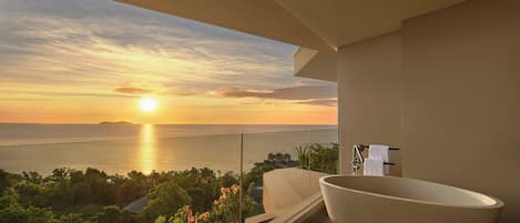A modern bathroom with a large bathtub, a shower, and a view of the ocean at sunset.