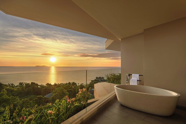 Un baño moderno con una bañera grande, ducha y vista al océano al atardecer.