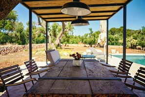 Mesa de jantar de madeira ao ar livre com bancos, luminária pendente e vista para uma piscina.