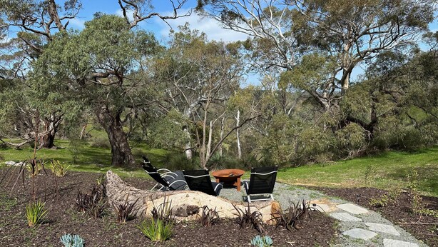 Uma área externa com mesas e cadeiras, rodeada por um jardim com diversas plantas e um caminho de pedra.
