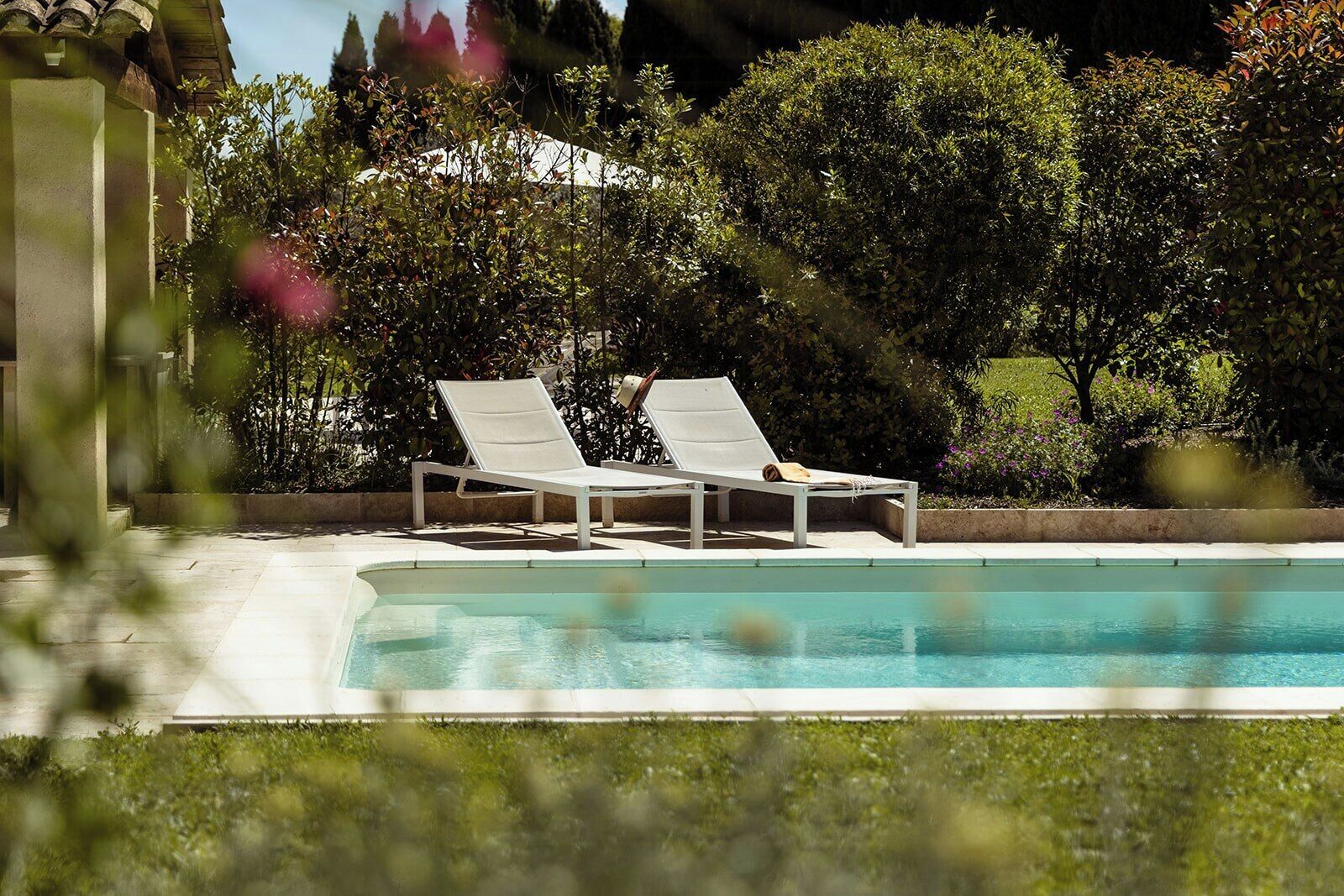 A swimming pool with two white lounge chairs, surrounded by greenery and a building in the background.