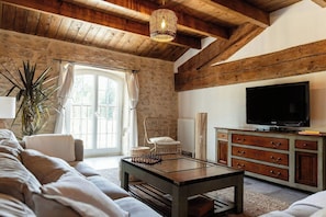 A cozy living room with a stone wall, wooden ceiling beams, a large window with curtains, a sofa, a coffee table, a television, and a cabinet. - Farmhouse with pool near Saint-Rémy-de-Provence (Mas Blanc des Alpilles)