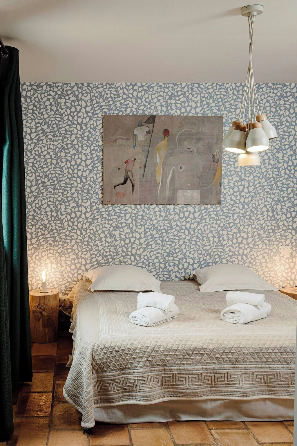A hotel room with a patterned wallpaper, a bed with a geometric-patterned bedspread, a painting on the wall, and a modern chandelier.