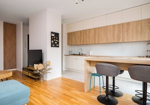 Una cocina moderna con armarios de madera, una zona de comedor con dos taburetes de bar, un televisor y piso de madera.