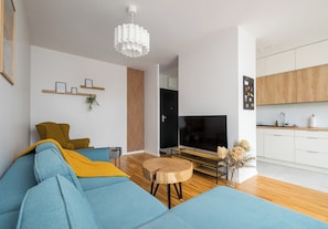 A modern living room with a sofa, TV, wooden floor, and a kitchen area in the background. - Fabryka na Pradze Charming Apartment (Warszawa)