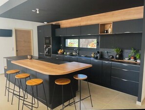 A modern kitchen with a central island, bar stools, dark cabinetry, and a window providing natural light. - Designer villa near the Gulf of Saint-Tropez (Grimaud)