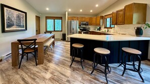 A modern kitchen with wooden cabinets, a large island with stools, a dining table with chairs, and a framed picture on the wall. - Experience Whitefish Retreat at Poplar Place, a 3-bedroom haven in scenic Whitefish, MT. (Whitefish)