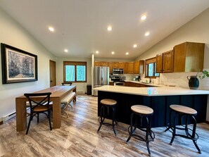 A modern kitchen with wooden cabinets, a large island with stools, a dining table with chairs, and a framed picture on the wall. - Experience Whitefish Retreat at Poplar Place, a 3-bedroom haven in scenic Whitefish, MT. (Whitefish)