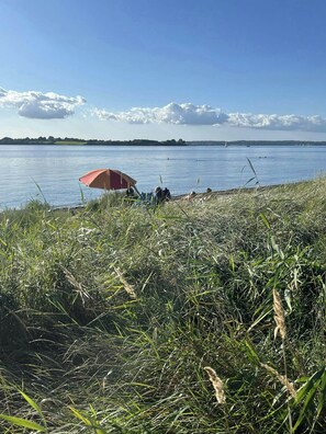 A grassy area with a red umbrella, a body of water, and a distant shoreline. - Sleep under the stars near Brunsnæs Beach (Broager)