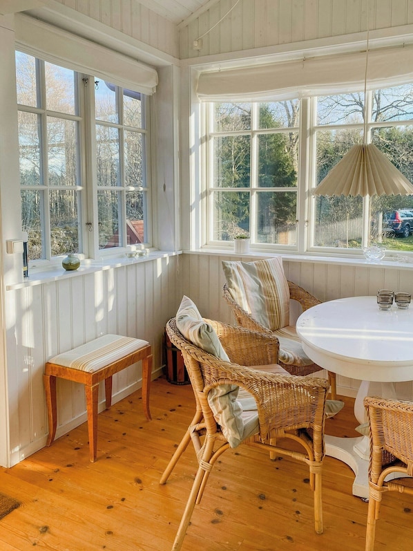 Ein Zimmer mit Holzboden, Korbstühlen, einem runden Tisch und großen Fenstern mit weißem Rahmen.
