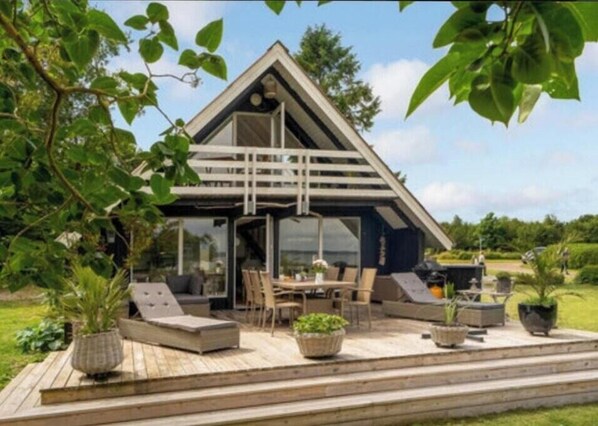 A modern house with a triangular roof, a deck with outdoor furniture, and a large window overlooking a garden. - Fjord views and spa by Limfjorden (Roslev)