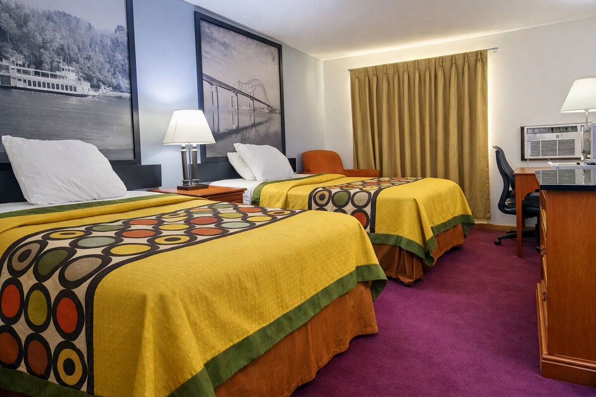 Habitación de hotel con dos camas, un escritorio, una silla, una lámpara, un cuadro de un puente y una ventana con cortinas.