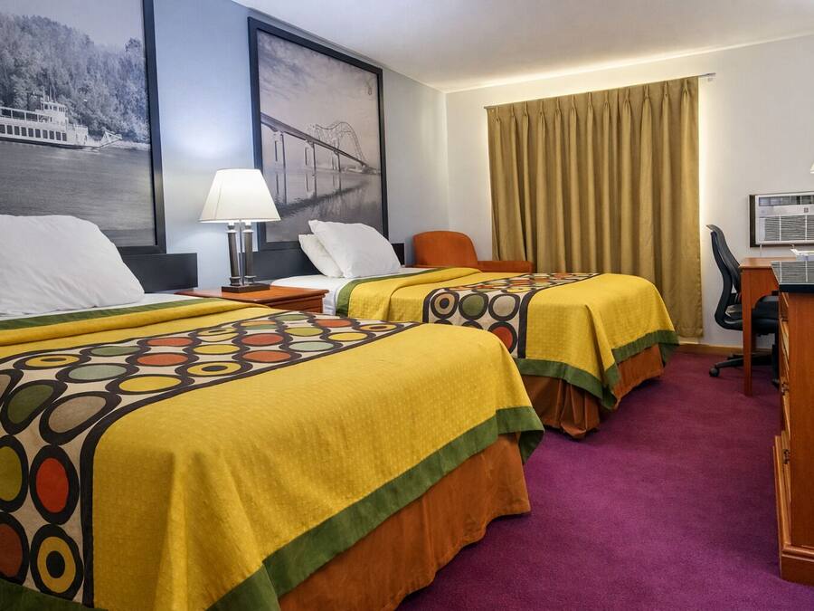 Habitación de hotel con dos camas, un escritorio, una silla, una lámpara, un cuadro de un puente y una ventana con cortinas.