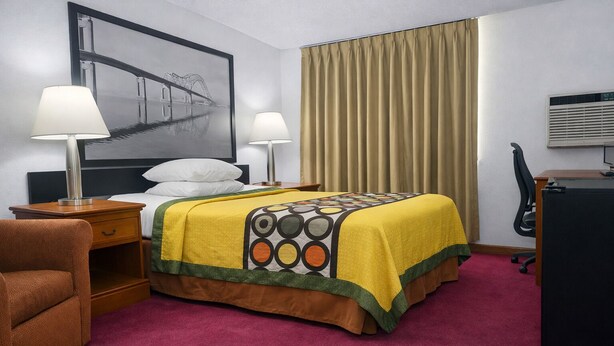 A hotel room with a bed, two bedside lamps, a desk with a computer, a chair, a large window with curtains, and a wall-mounted air conditioning unit.