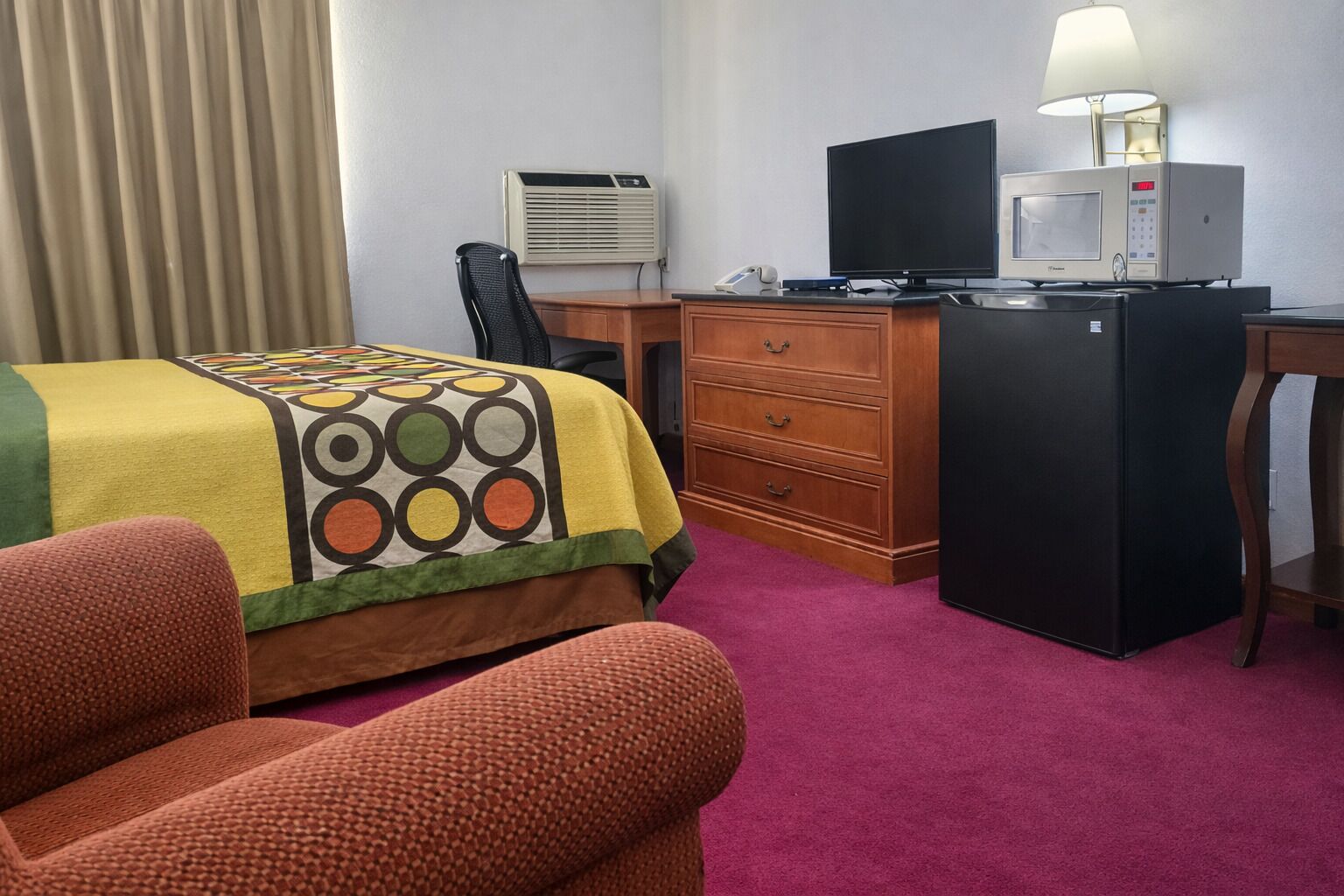Ein Hotelzimmer mit einem Bett, einem Schreibtisch mit Computer, einem Kühlschrank, einer Klimaanlage und einer Lampe.