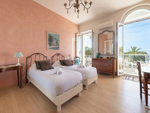 A bedroom with two beds, a dresser, a desk with a lamp, a painting, and a balcony with a view. - 2-Br Bourgeois Apartment – Croisette & Sea View (Cannes)