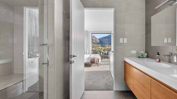 A modern bathroom with a shower, a large mirror, and a view of a bedroom with a bed and a view of mountains.