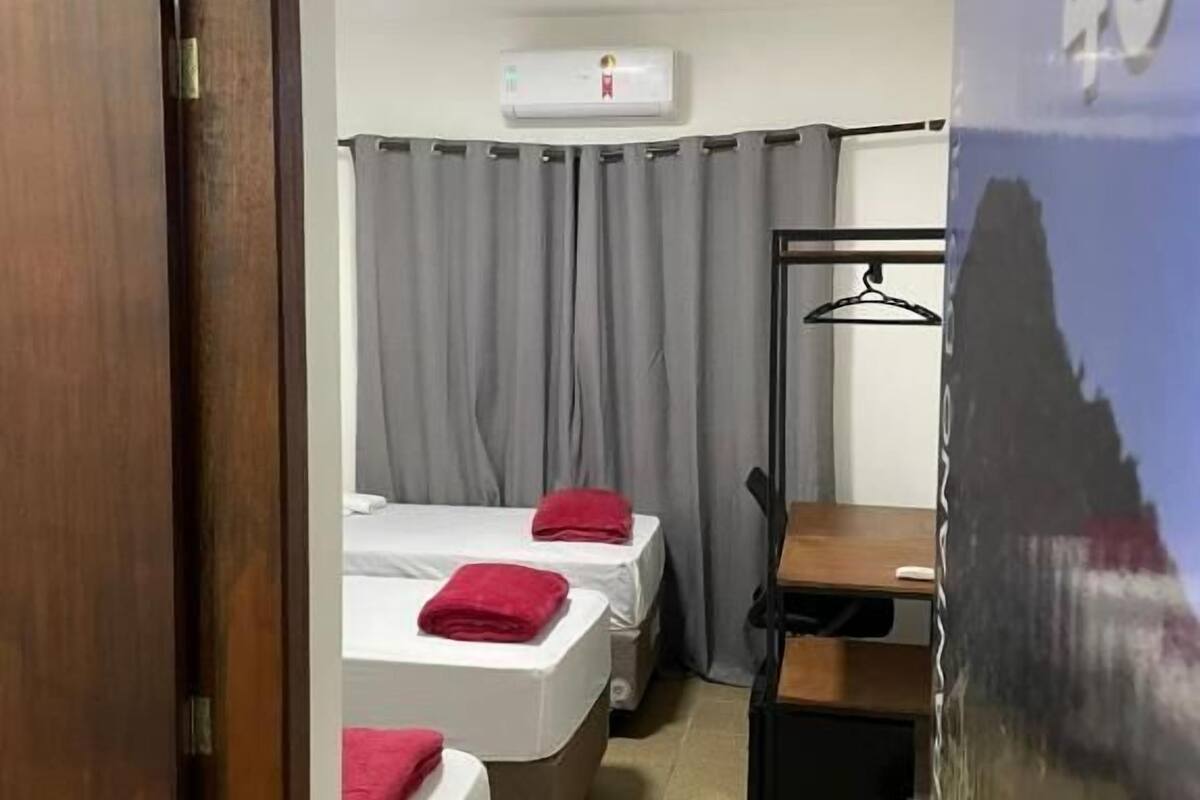 Habitación de hotel con dos camas, cortinas grises, un escritorio de madera, un perchero negro y un aire acondicionado instalado en la pared.