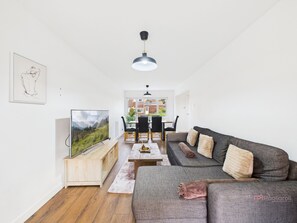 A modern living room with a flat-screen TV, a gray sofa, a wooden cabinet, and a dining area with a table and chairs.