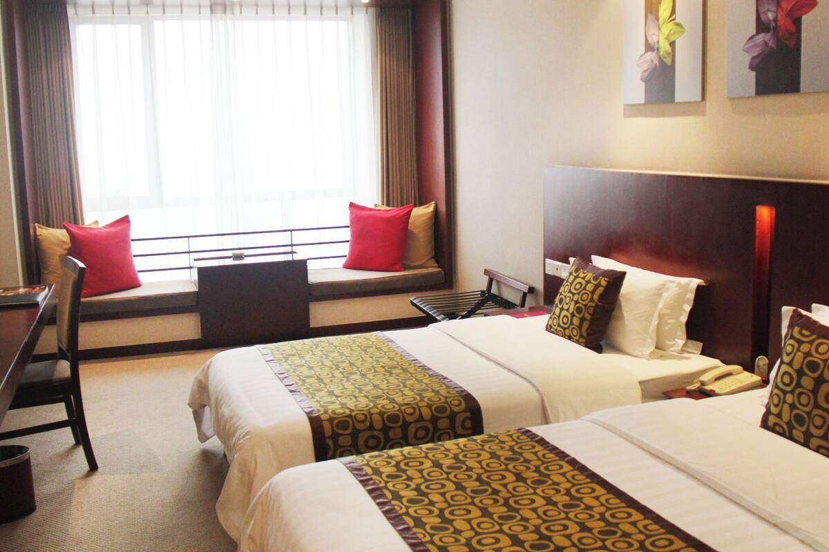 A hotel room with two beds, a desk, a chair, a window with curtains, and artwork on the wall.
