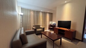 A hotel room with a sofa, a coffee table, a TV on a wooden stand, a desk with a chair, and a large window with curtains. - Beijing Wanshang Garden Hotel (Beijing)