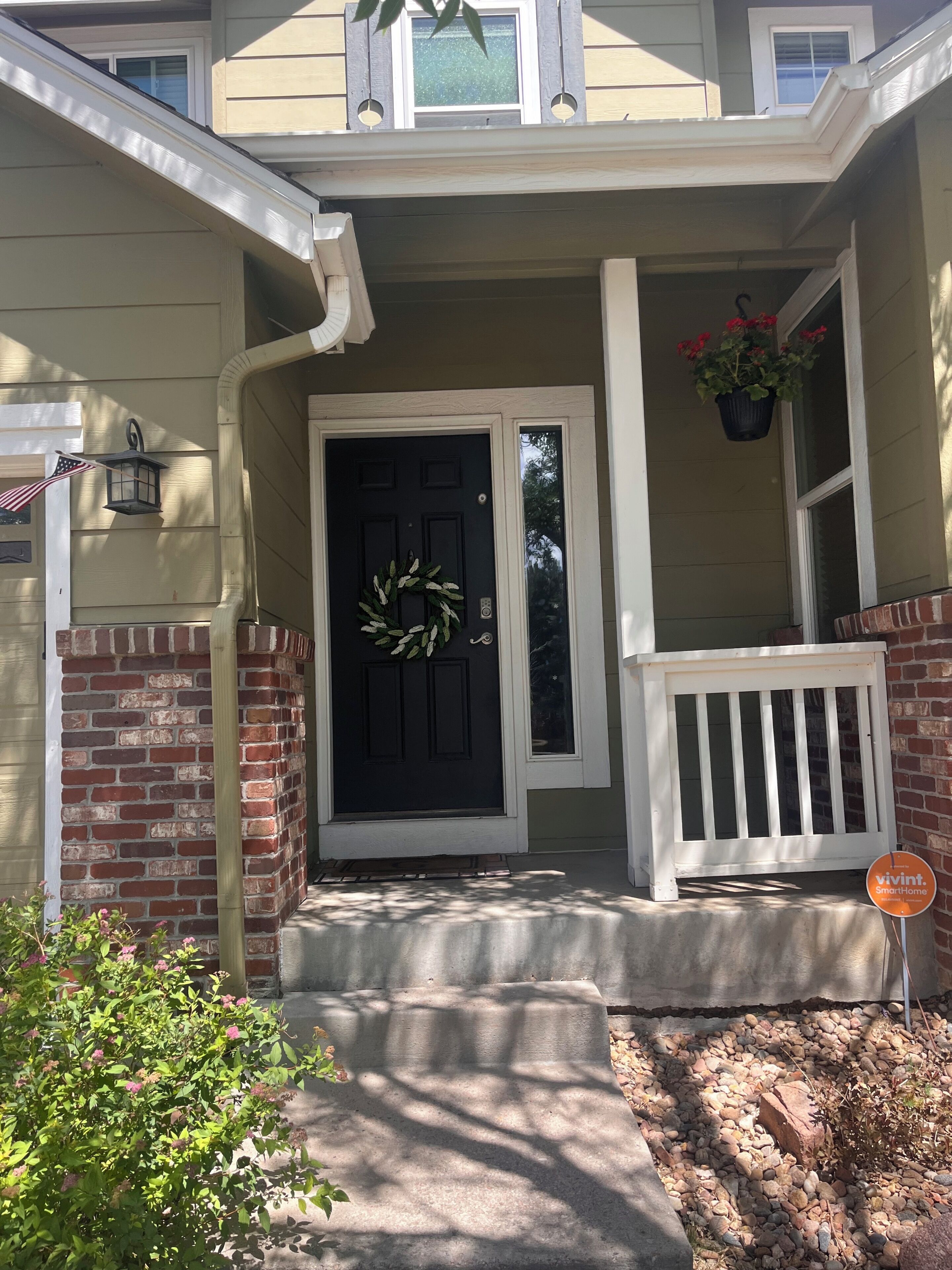 A house with a black front door, a brick wall, a porch with a white railing, and a hanging flower pot.