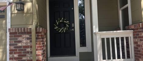 A house with a black front door, a brick wall, a porch with a white railing, and a hanging flower pot.
