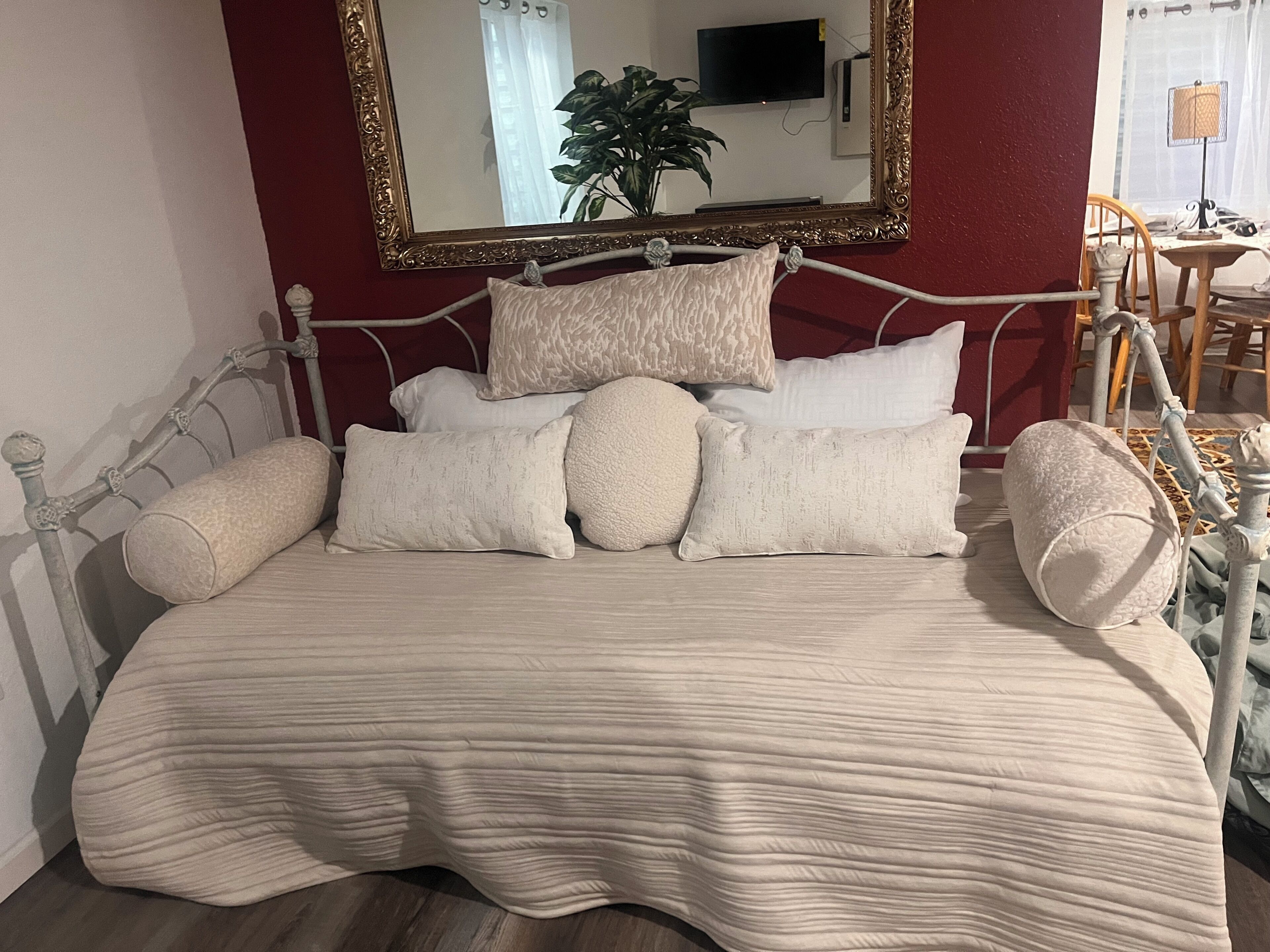 Ein Bett mit einer strukturierten, beigefarbenen Tagesdecke, mehreren Kissen, einem Metall-Kopfstück und einem Spiegel mit aufwendigem Rahmen.