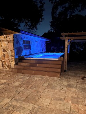 Une façade en pierre et une terrasse en bois menant à un espace équipé d’un jacuzzi, le tout abrité par une pergola.