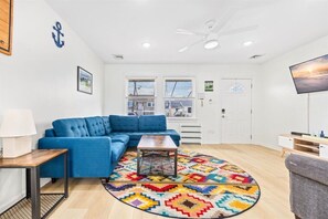 A living room with a blue sofa, a coffee table, a colorful rug, a ceiling fan, and a TV on the wall. - Ventnor Home | BBQ + Fenced Yard | Beach Gear Incl (Ventnor City)