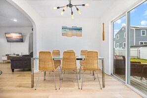 A dining area with a glass table, wicker chairs, a modern chandelier, and a large window offering a view of the outside. - Ventnor Home | BBQ + Fenced Yard | Beach Gear Incl (Ventnor City)