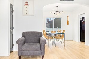 A modern living room with a grey armchair, a dining table with wicker chairs, a chandelier, and a black refrigerator. - Ventnor Home | BBQ + Fenced Yard | Beach Gear Incl (Ventnor City)