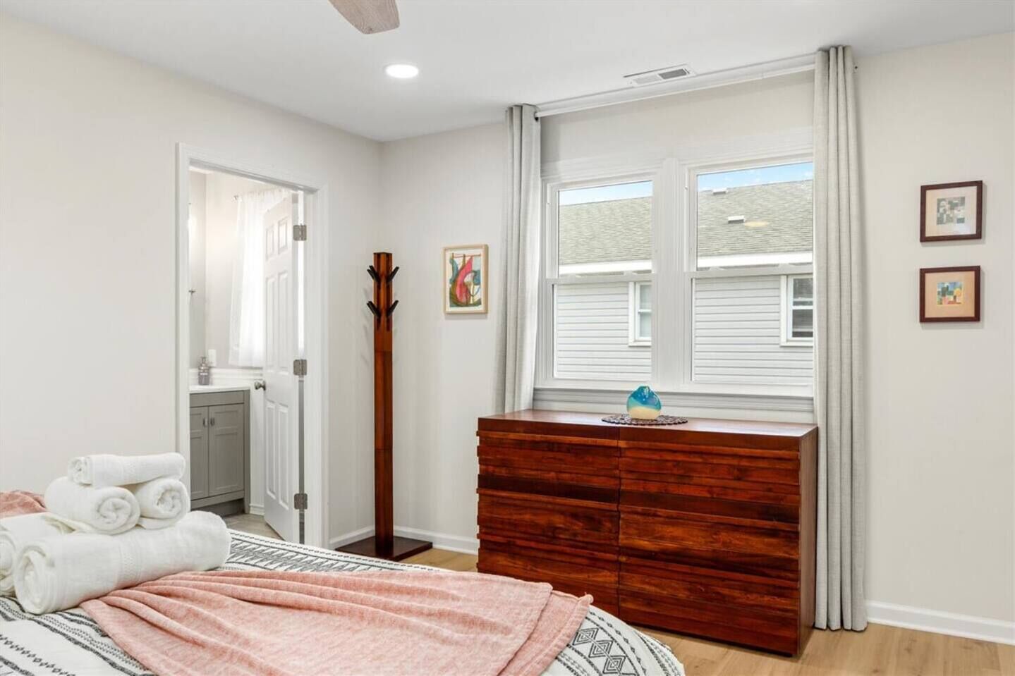Un dormitorio con cama, cómoda, perchero, ventana con cortinas y puerta que da a otra habitación.