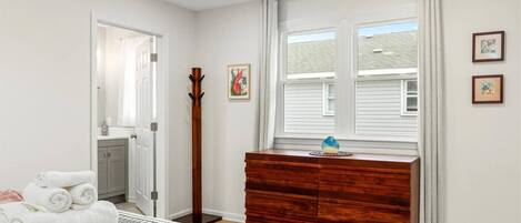 Un dormitorio con cama, cómoda, perchero, ventana con cortinas y puerta que da a otra habitación.