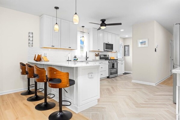 A modern kitchen with white cabinets, a central island with bar stools, a microwave, oven, and a ceiling fan. - Modern Ventnor Retreat | Sleeps 8 | Backyard (Ventnor City)