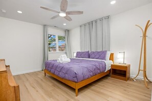 A bedroom with a wooden bed, a nightstand, a lamp, a ceiling fan, and a window with curtains. - Modern Ventnor Retreat | Sleeps 8 | Backyard (Ventnor City)