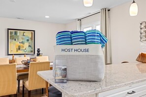 A modern kitchen with a marble countertop, a dining area with wooden chairs, and a striped pillow on a white cabinet labeled 'HOST HOUSE'. - Modern Ventnor Retreat | Sleeps 8 | Backyard (Ventnor City)