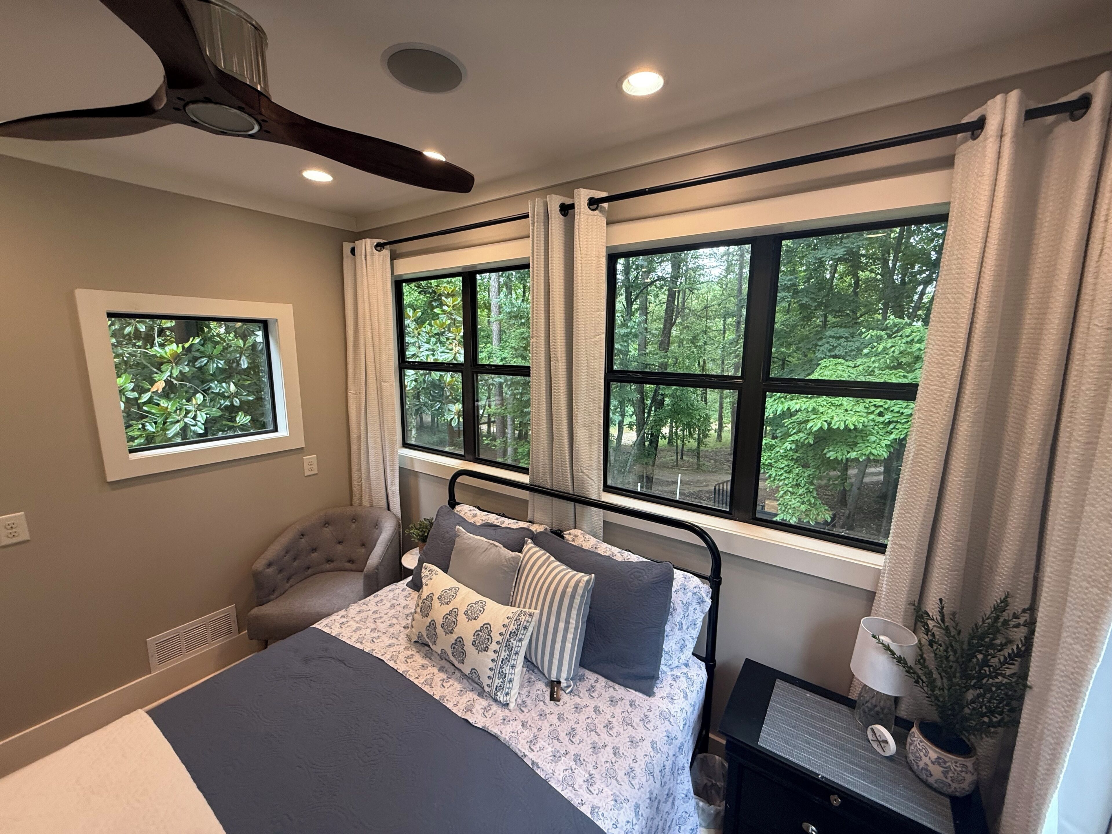 Une chambre à coucher comprenant un lit, une chaise, une table de chevet, un ventilateur de plafond et des fenêtres donnant sur la forêt.