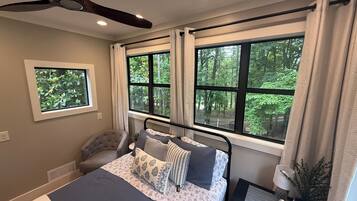 Une chambre à coucher comprenant un lit, une chaise, une table de chevet, un ventilateur de plafond et des fenêtres donnant sur la forêt.