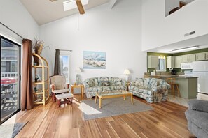 A living room with a sofa, armchairs, a coffee table, a bookshelf, and a painting on the wall. - Bright Condo w/ Loft for 8! Bay Tree 24B (Little River)