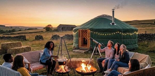 A yurt with a telescope, hay bales, and a fire pit in a rural setting.