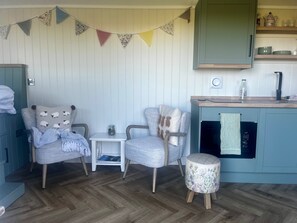A cozy room with two armchairs, a small table, a decorative stool, a kitchen area with a counter, and a wall-mounted shelf.