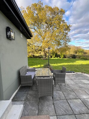 Eine Terrasse mit Korbmöbeln und einem Tisch, einem Laternenpfosten und Blick auf einen von Bäumen gesäumten Garten.
