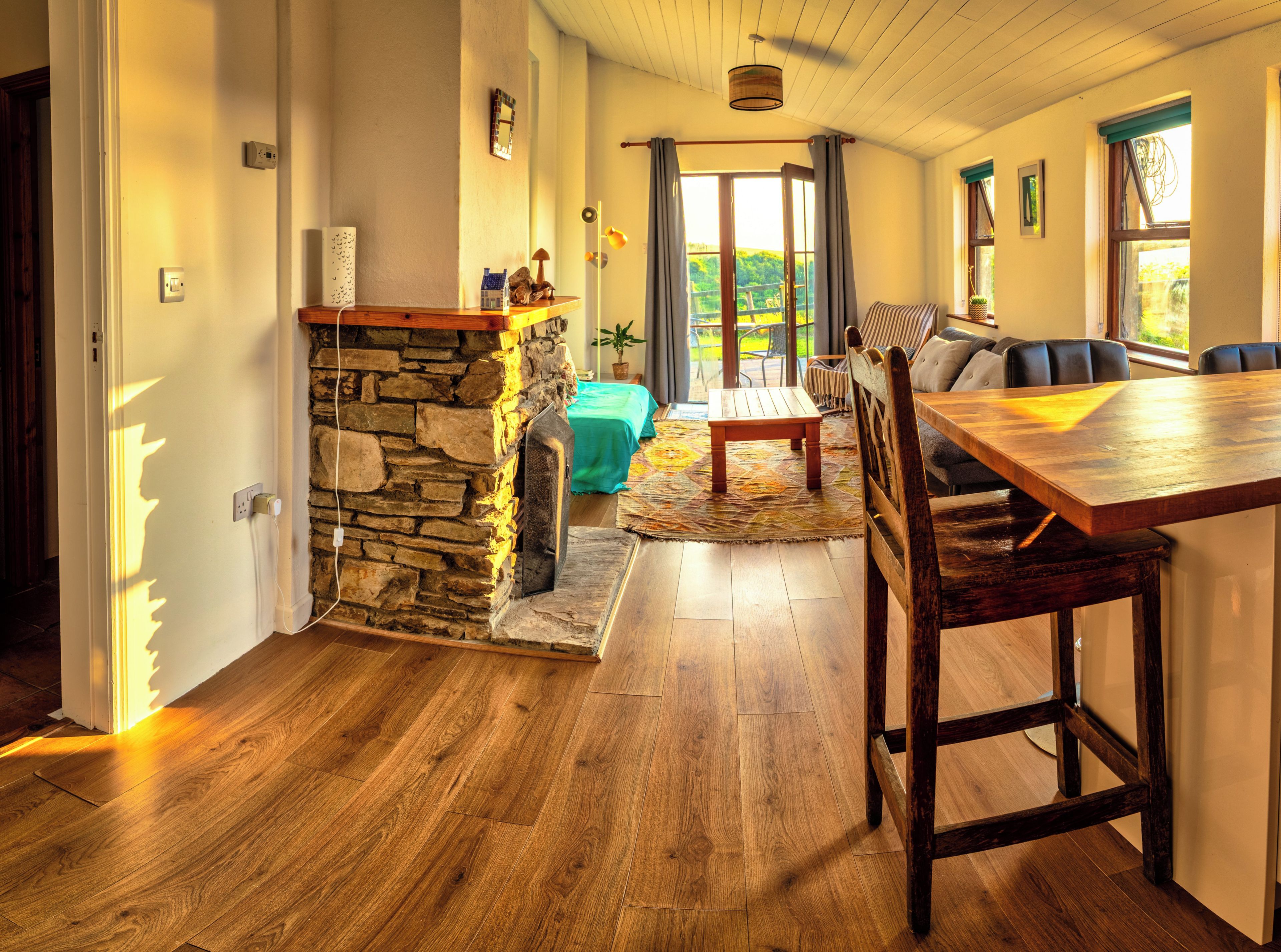 A room with a stone wall, wooden flooring, a dining table with chairs, a sofa, and a view through a large window.