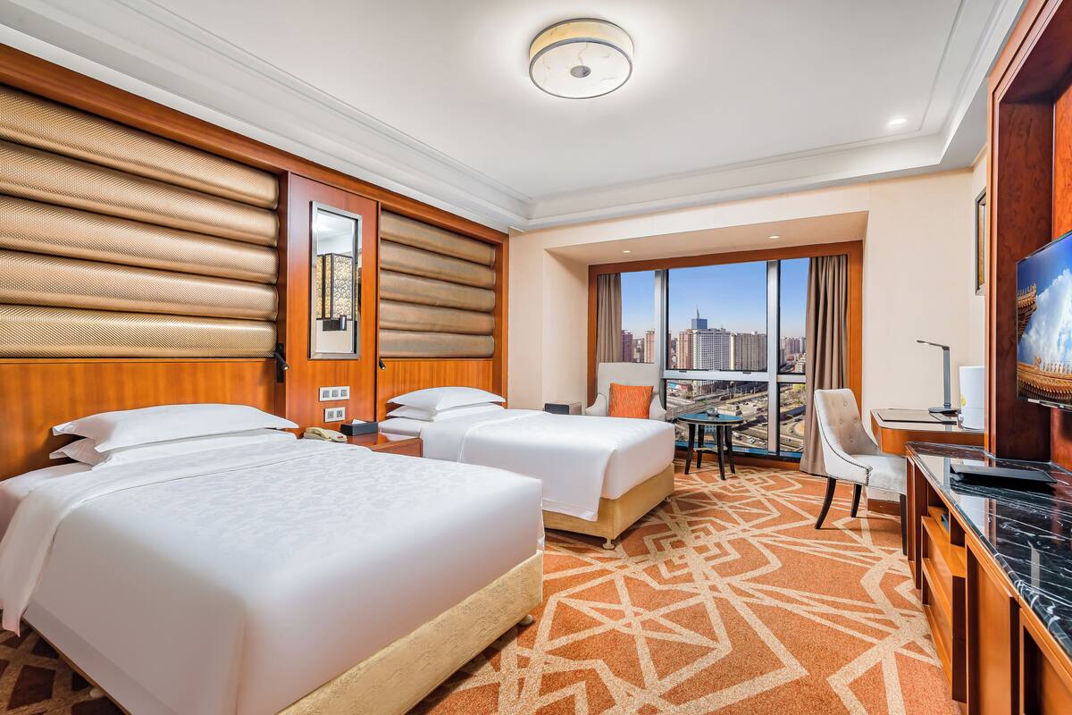 Habitación de hotel con dos camas, un escritorio, una silla, un televisor y un ventanal con vistas a la ciudad.