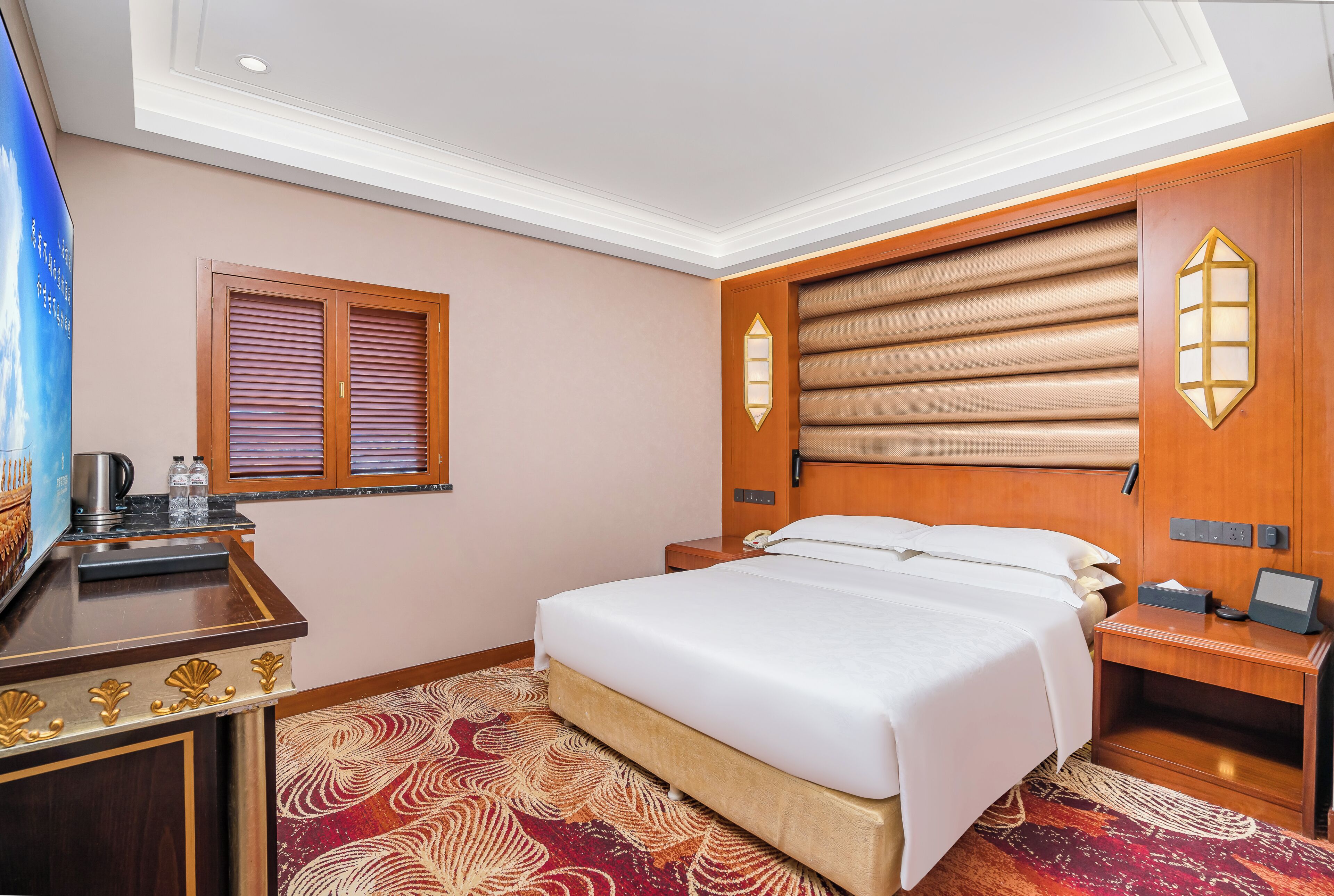 A hotel room with a bed, a desk with a lamp, a window with blinds, and a wall-mounted light fixture.