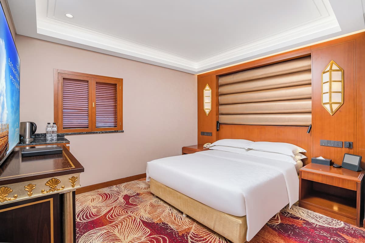 A hotel room with a bed, a desk with a lamp, a window with blinds, and a wall-mounted light fixture.