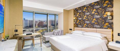 Habitación de hotel con una cama grande, un escritorio, una silla y vistas de la ciudad a través de amplios ventanales.
