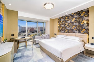 Ein Hotelzimmer mit einem großen Bett, einem Schreibtisch, einem Sessel und Blick auf die Skyline durch große Fenster.